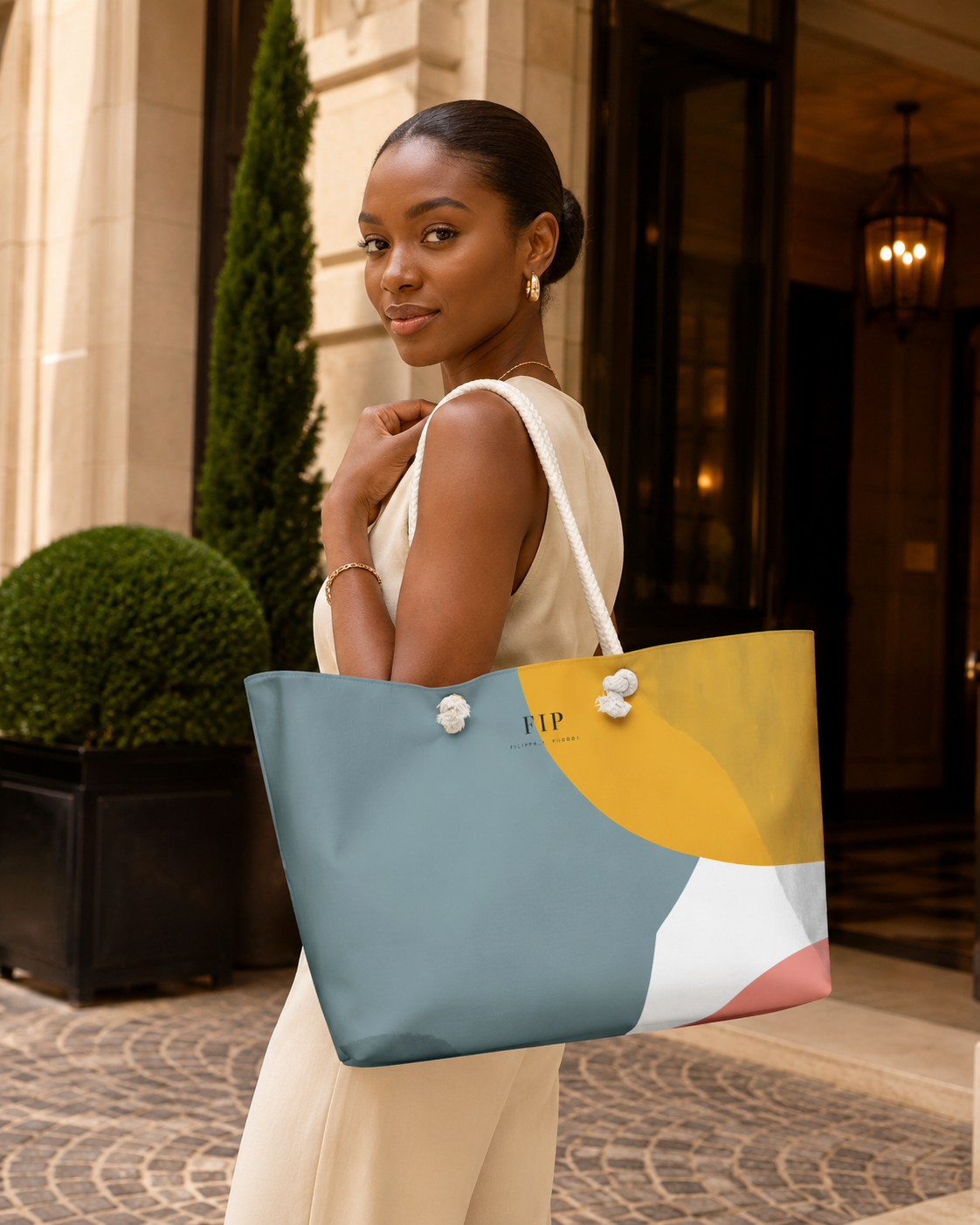 cream-colored dress carries a large, colorful tote bag with white rope handles. The bag features a modern geometric color-blocked design in slate blue, mustard yellow, white, and soft pink,