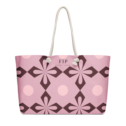 A pink tote bag with a geometric floral pattern in dark brown and light pink, featuring cream-colored rope handles. 